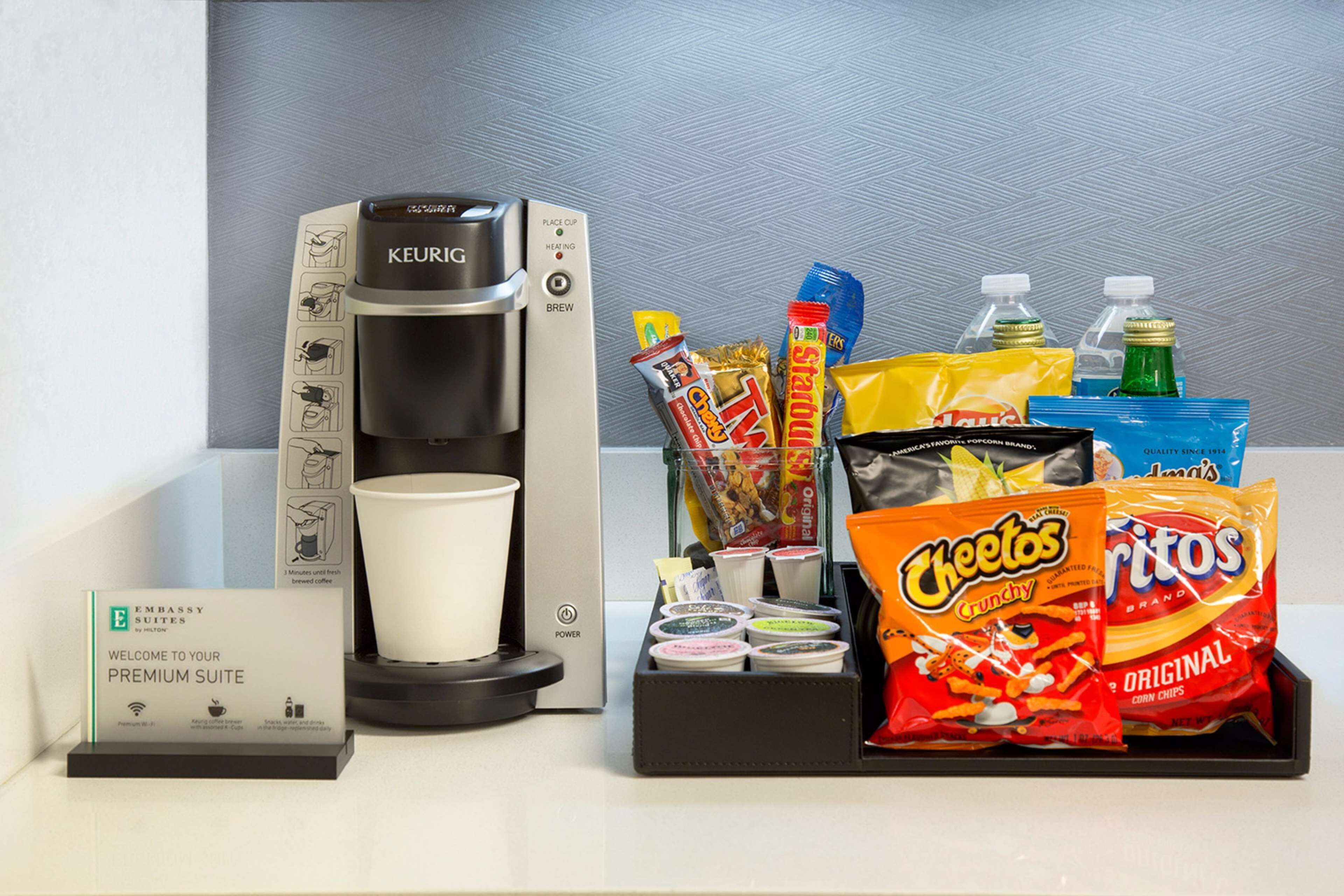 Una cafetera, una taza y diversos refrigerios, como papas fritas y agua embotellada.