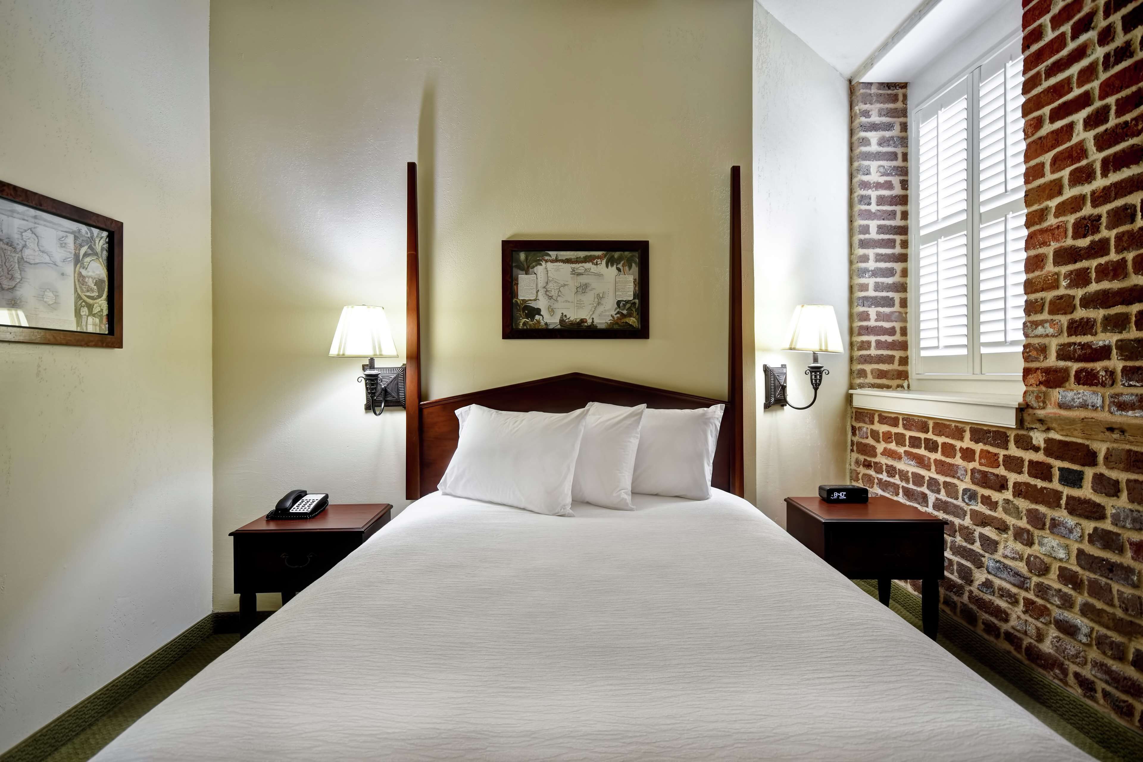 A hotel room with a large bed, two bedside tables with lamps, a wooden headboard, a framed picture on the wall, and a brick wall on one side.