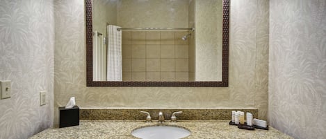 Een badkamer met een wastafel met granieten blad, een spiegel met houten lijst, en een douche zichtbaar op de achtergrond.