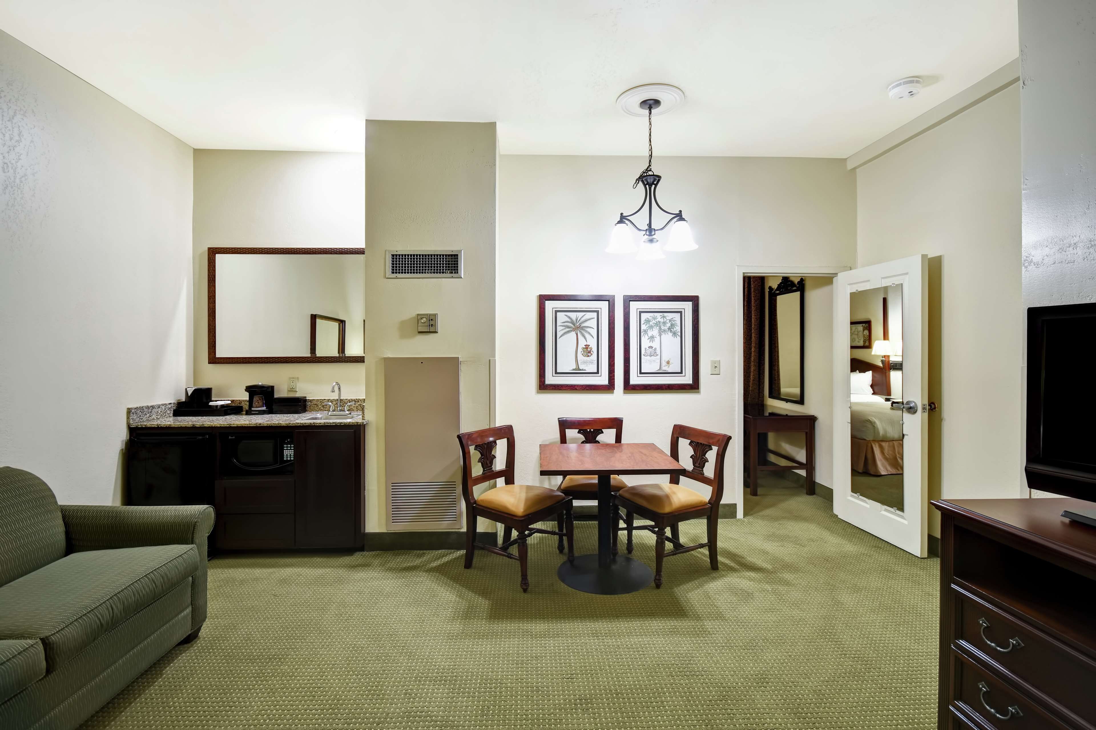 A compact hotel room with a kitchenette, a dining area with a table and chairs, a sofa, a TV, and a mirror.