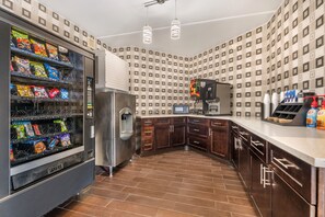 A kitchen with a vending machine stocked with snacks, a stainless steel refrigerator, a microwave, and a coffee machine.