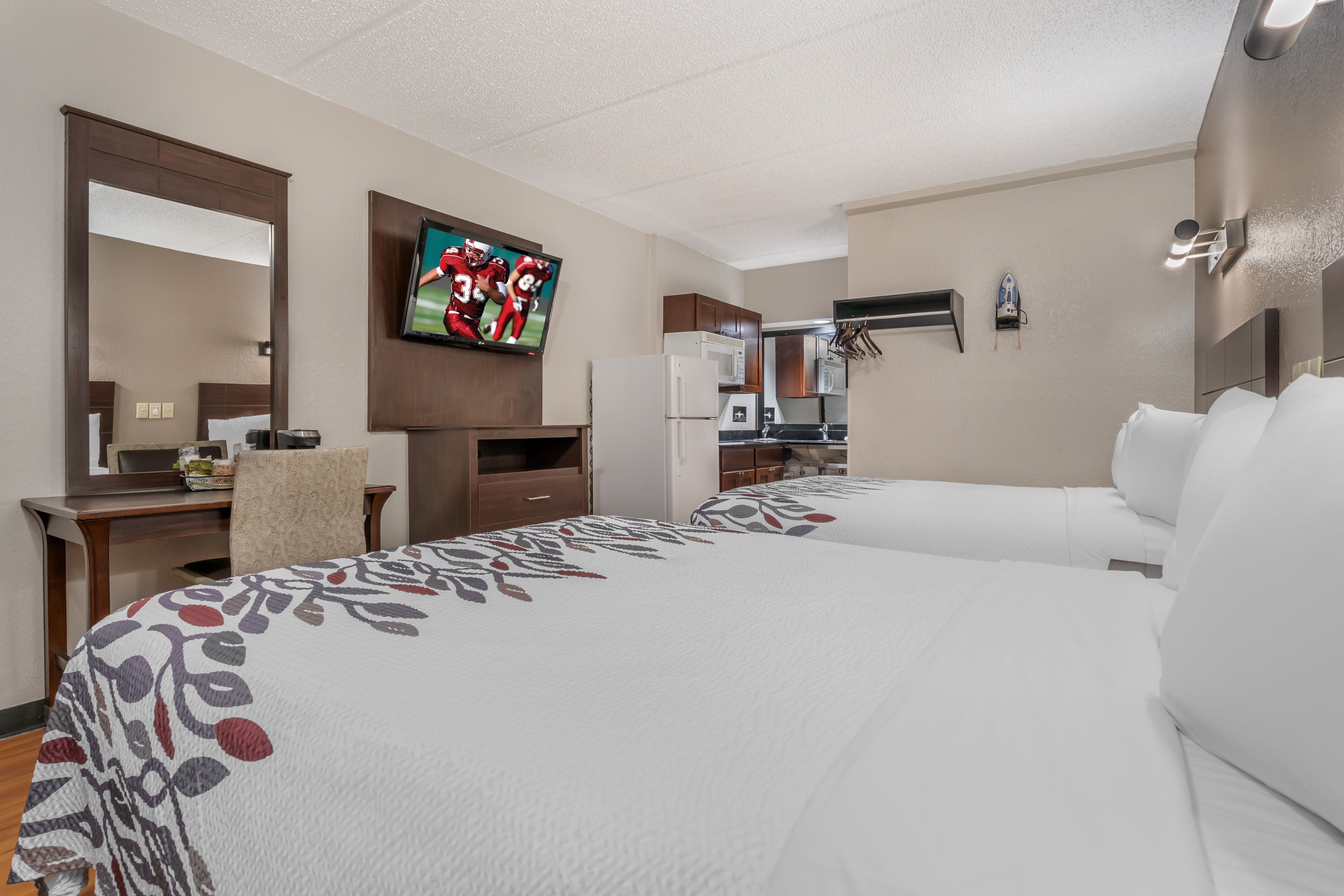 A hotel room with two beds, a TV, a desk, and a kitchenette.