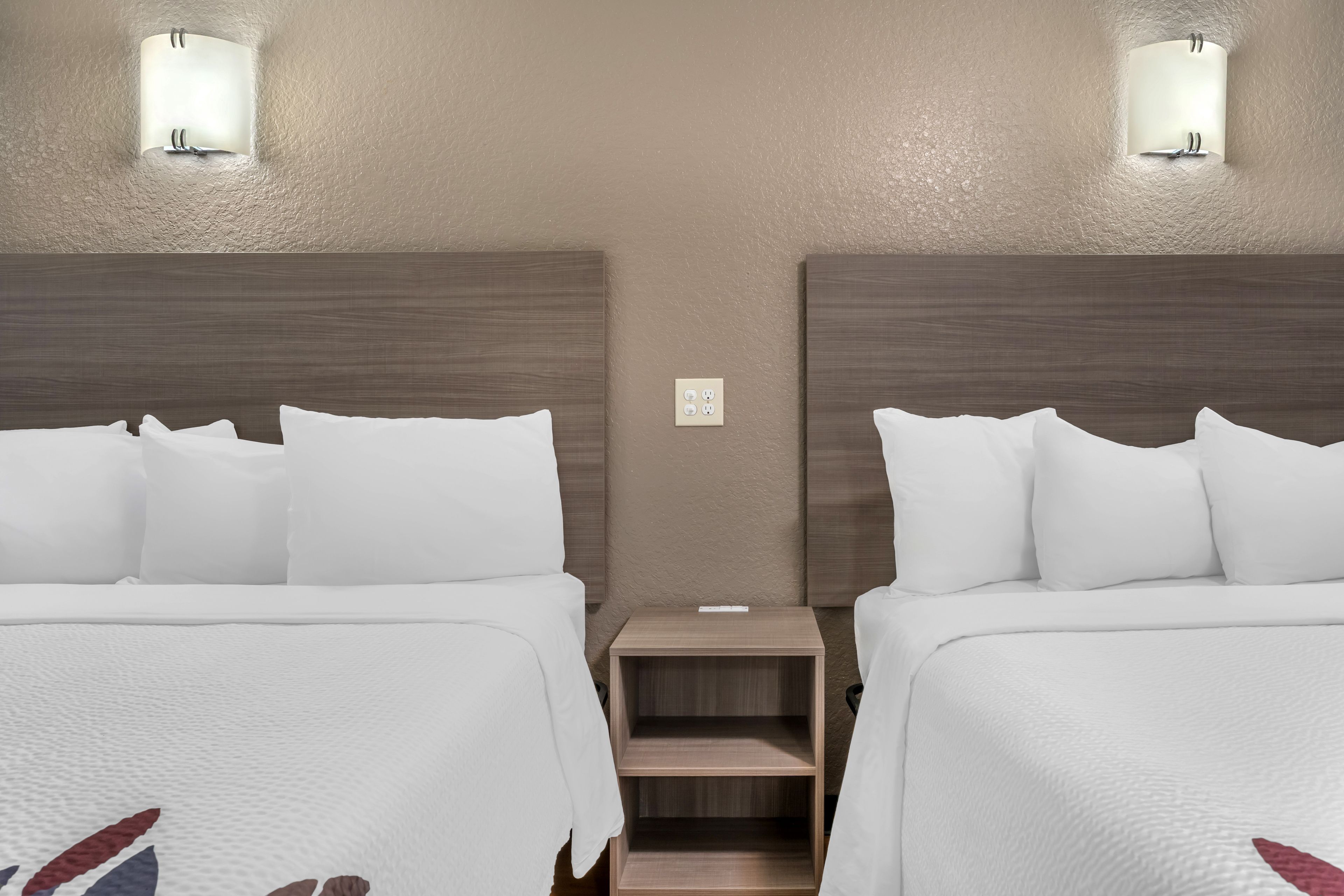 A hotel room with two beds, each with white bedding and pillows, a wooden headboard, and a small wooden nightstand between them.
