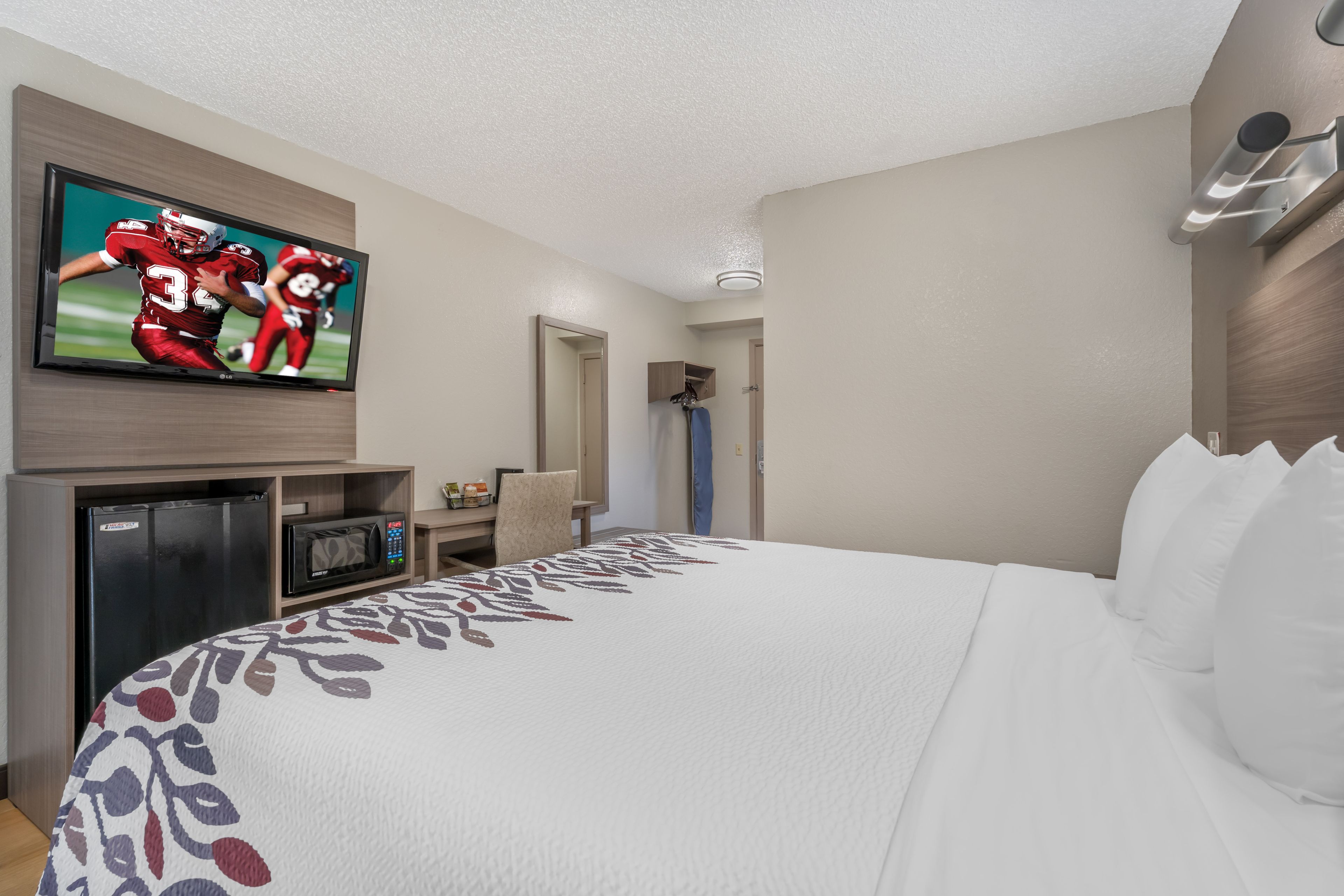 A hotel room with a large bed, a flat-screen TV mounted on the wall, a microwave, a refrigerator, and a small desk area.