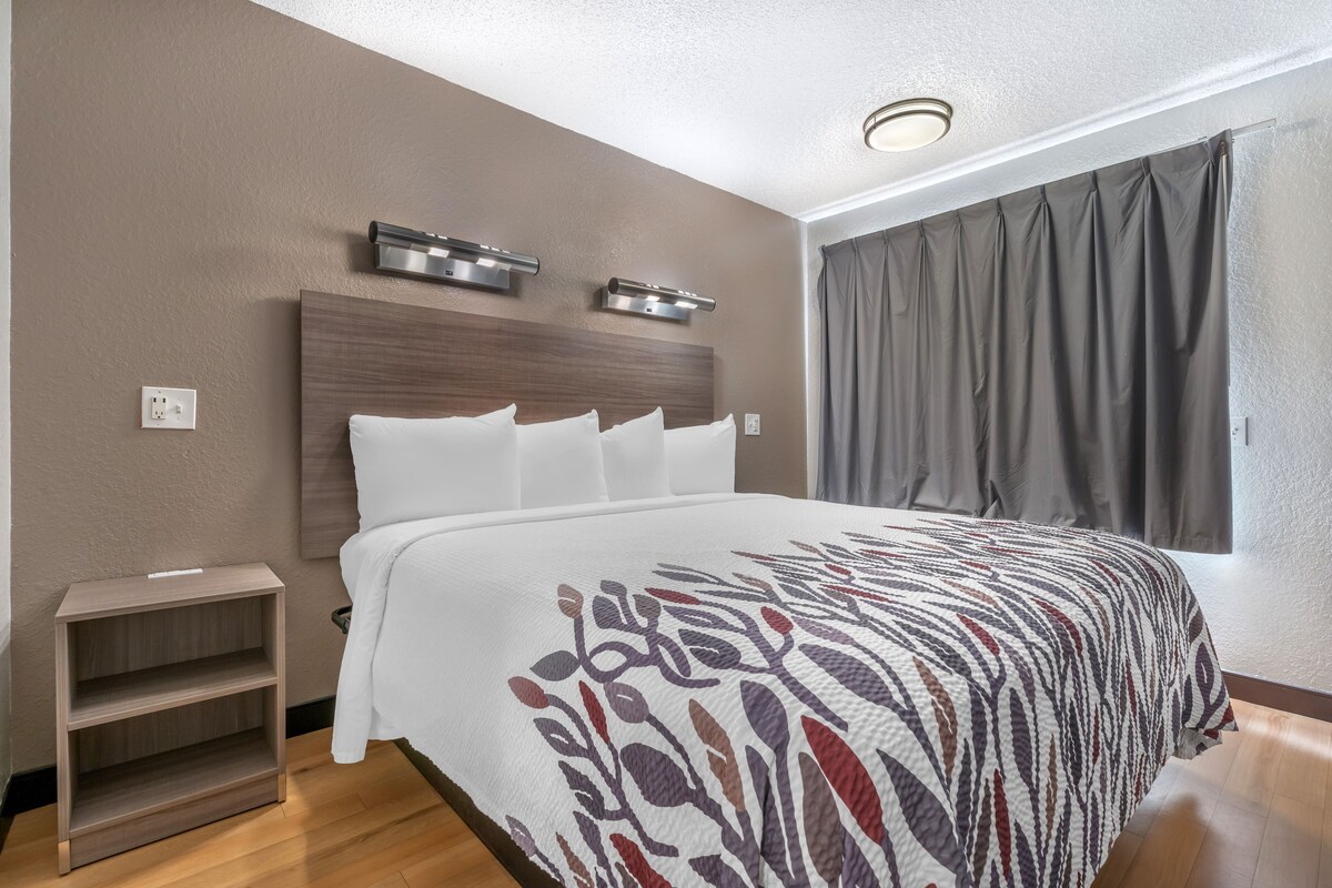 A hotel room with a large bed, a bedside table, a nightstand, a headboard with built-in lighting, and a window with curtains.