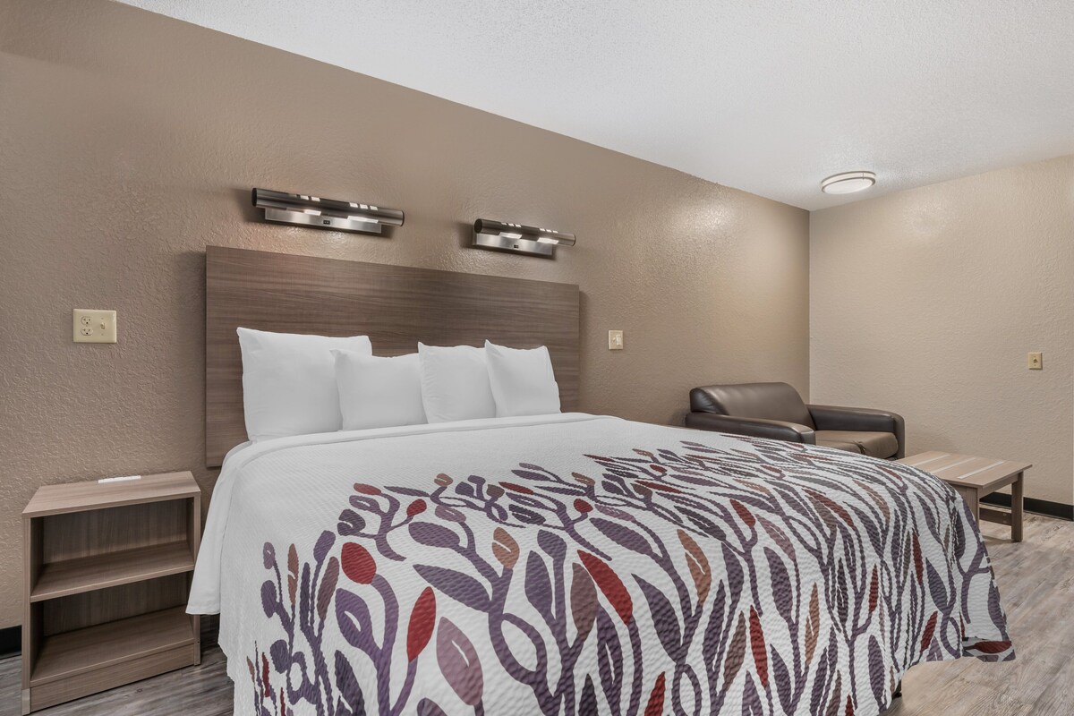 A hotel room with a large bed, a headboard, two wall-mounted lights, a wooden nightstand, a chair, and a small table.