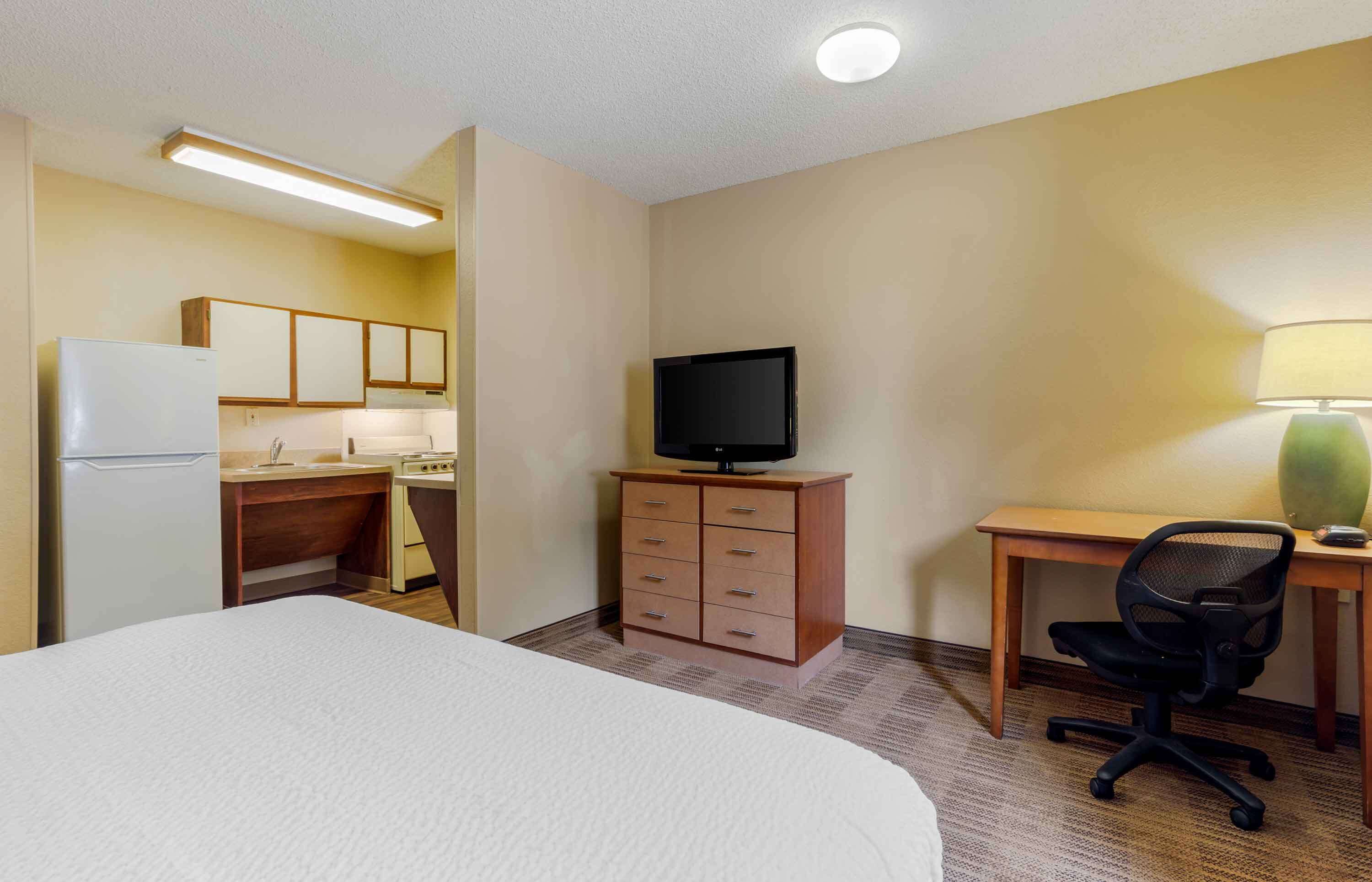 A hotel room with a bed, a desk with a chair, a television, a dresser, and a kitchenette.