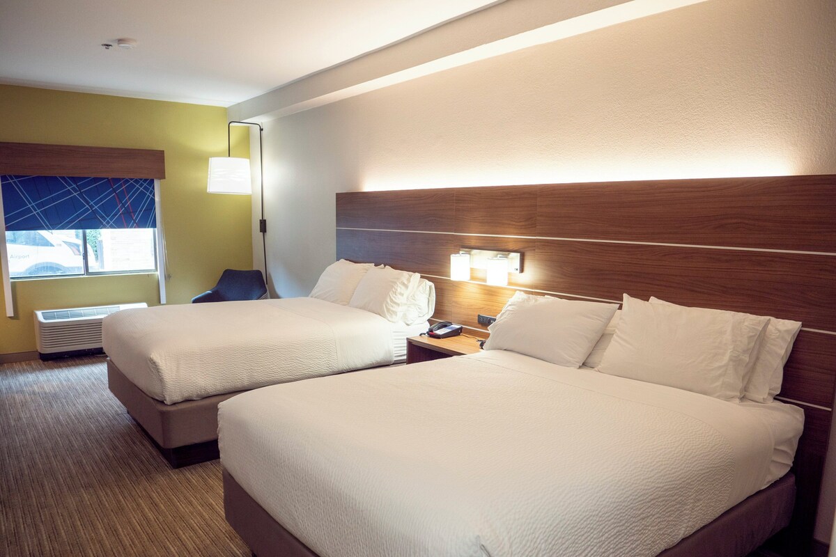 A hotel room with two beds, a window with a view, a desk with a lamp, and an air conditioning unit.