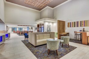 A spacious lobby with a high ceiling, a seating area with a sofa and chairs, a reception desk, and a colorful wall art piece. - Holiday Inn Express & Suites Greenville Airport by IHG (Greer)