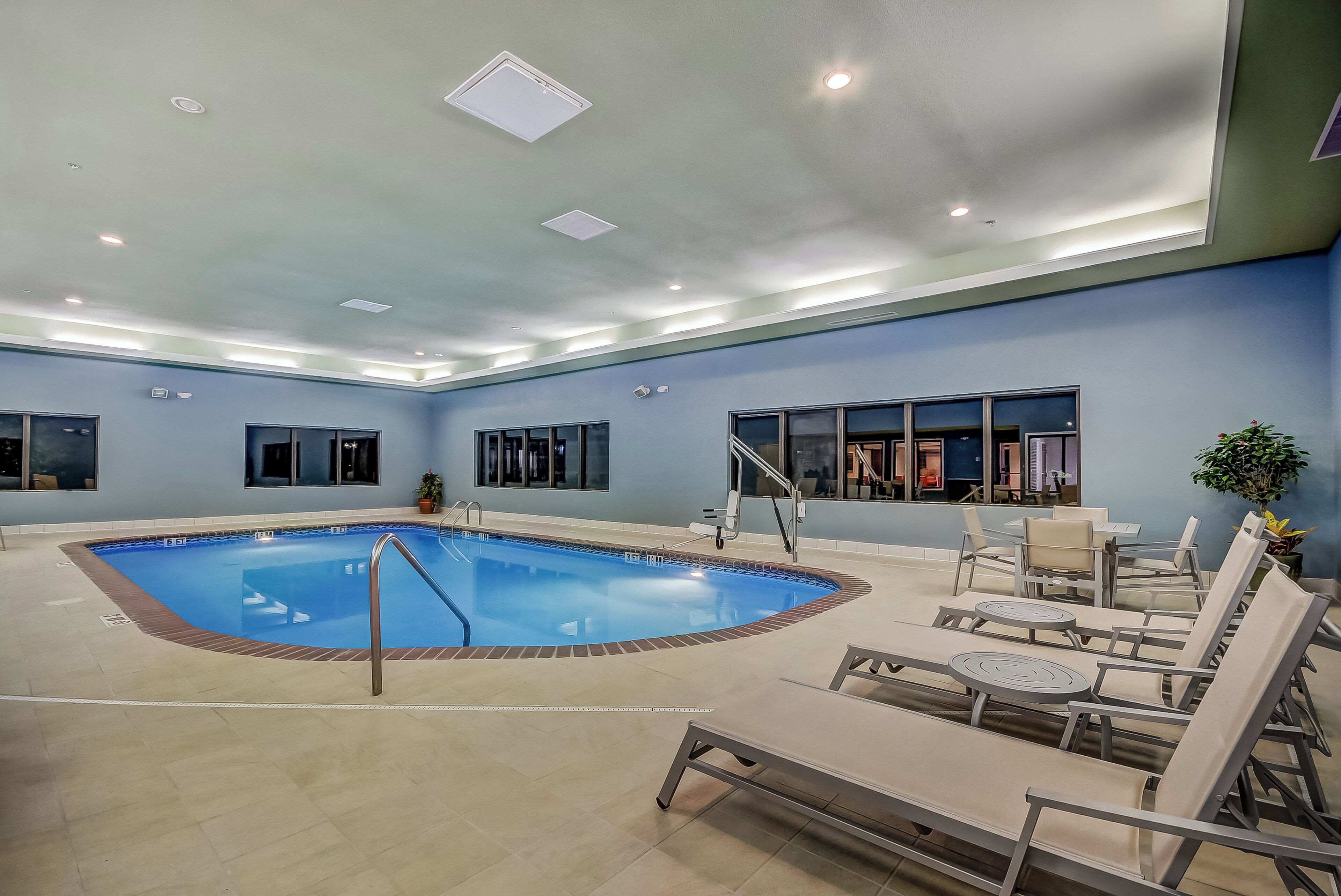 An indoor swimming pool with a tiled floor, surrounded by lounge chairs and tables, and a ceiling with recessed lighting.