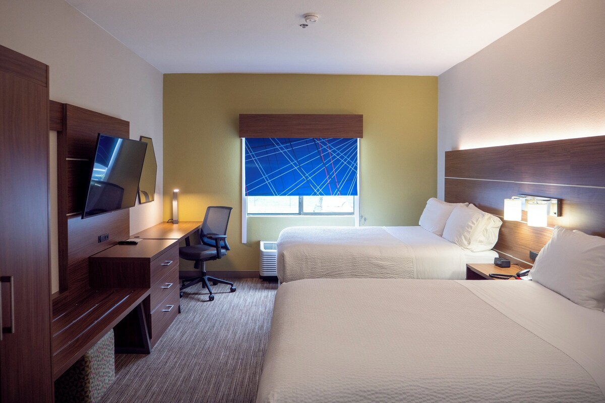 A hotel room with a bed, a desk with a chair, a television, and a window with a blue geometric pattern.