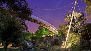 A suspension bridge over a river with a waterfall, surrounded by trees and rocks. - Holiday Inn Express & Suites Greenville Airport by IHG (Greer)