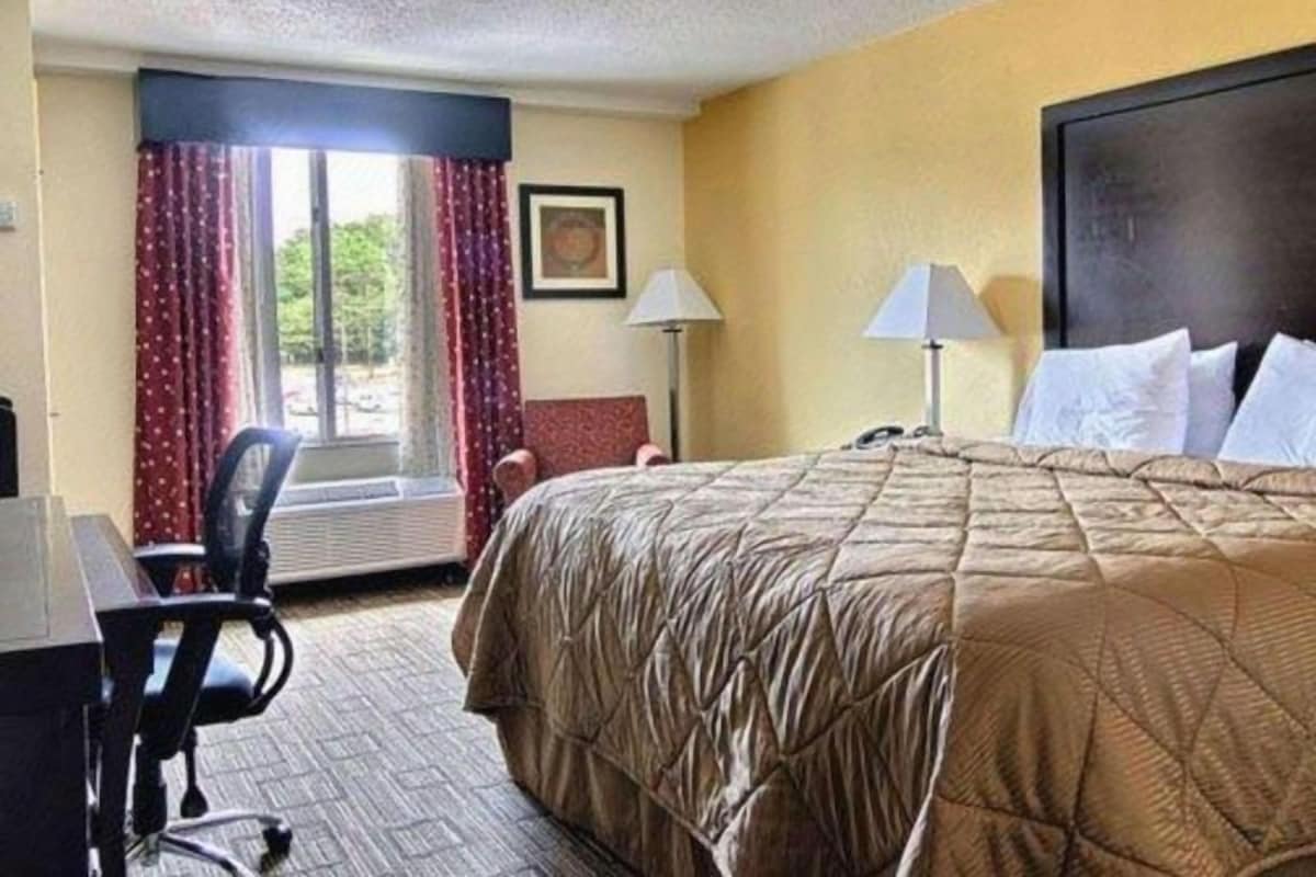 A hotel room with a bed, a desk, a chair, a window with curtains, and a lamp.