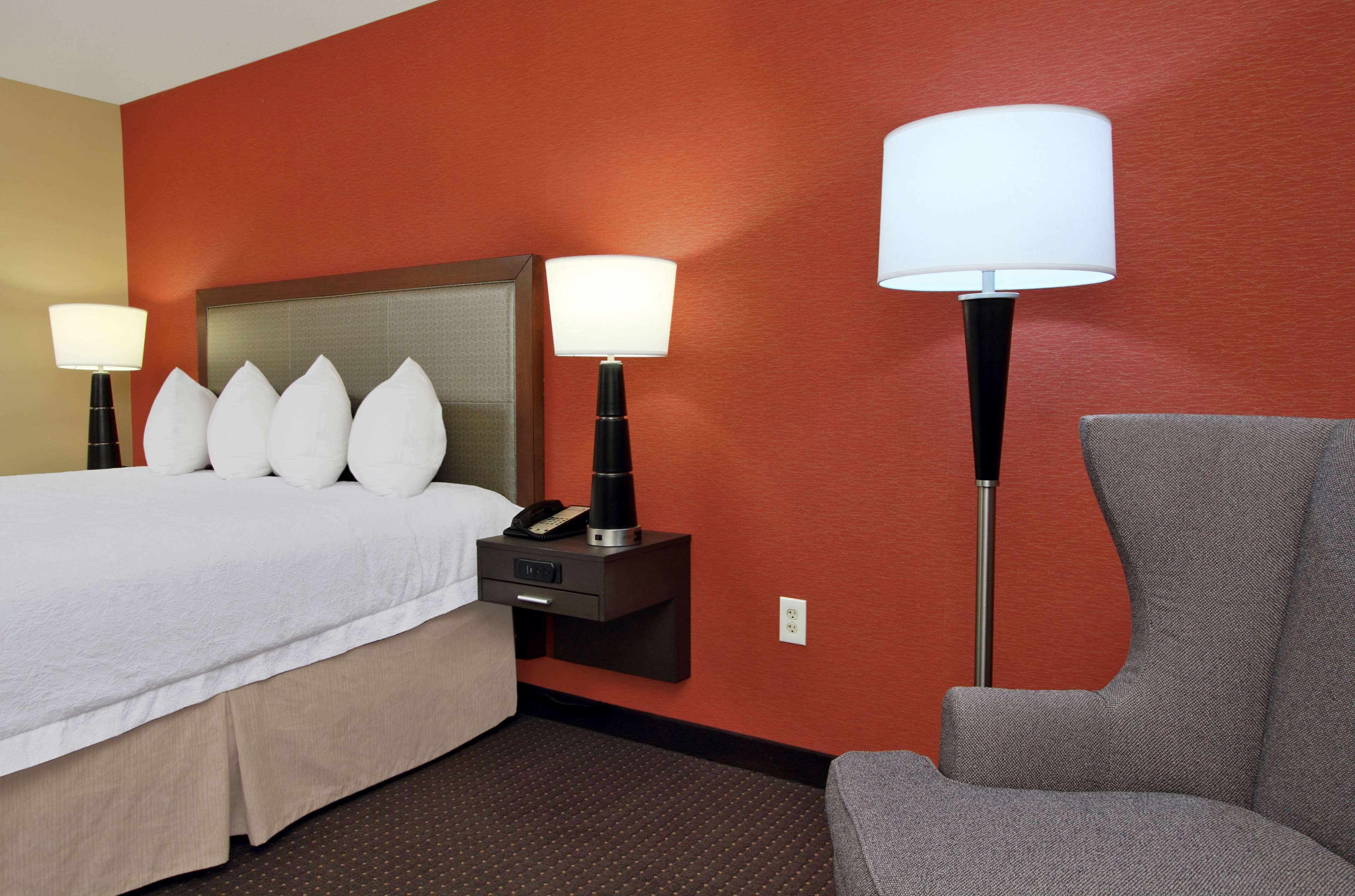 A hotel room with a bed, two bedside tables with lamps, a chair, and a red wall.
