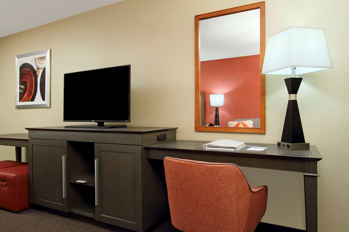 A hotel room with a desk, television, lamp, and a red chair.