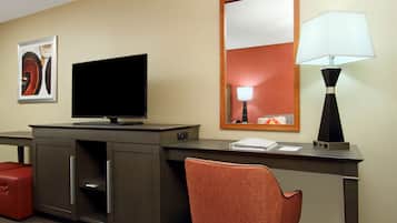 A hotel room with a desk, television, lamp, and a red chair.