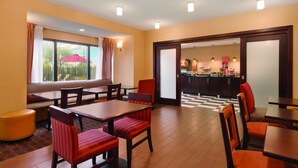 A hotel dining area with tables and chairs, a kitchen in the background, and a view through a window.