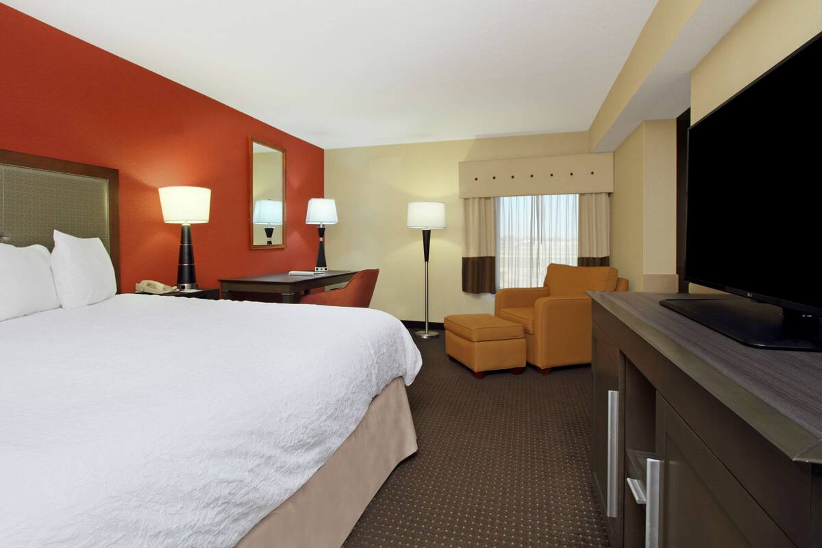 A hotel room with a large bed, a desk with a lamp, a TV, and a seating area with two chairs.