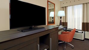 A hotel room with a large flat-screen TV on a wooden desk, a chair, a lamp, and a window with curtains.