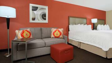 Habitación de hotel con un sofá gris, un reposapiés rojo, una cama con ropa de cama blanca y una lámpara de pie.