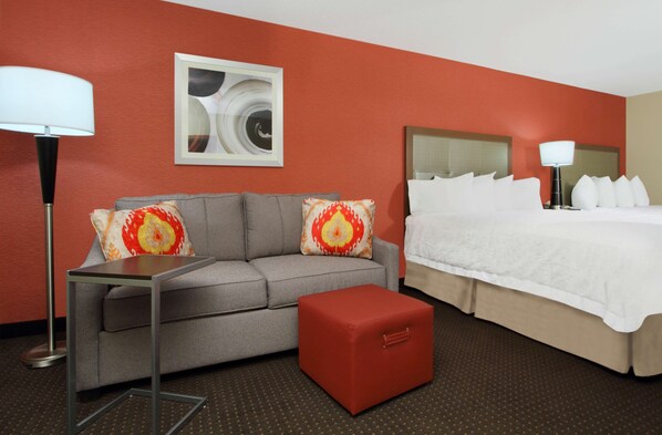 A hotel room with a grey sofa, a red ottoman, a bed with white bedding, and a floor lamp.