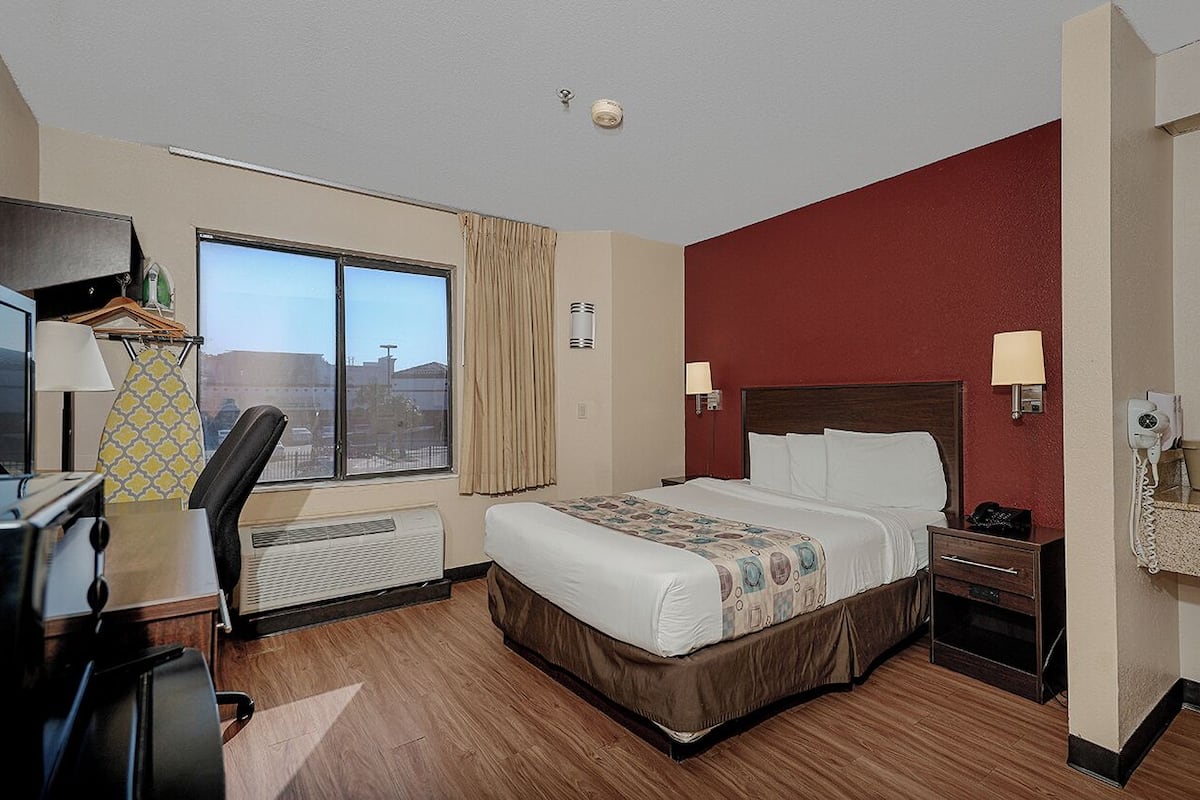 A hotel room with a large bed, a desk with a chair, a window with curtains, a red accent wall, and a bathroom with a mirror and sink.