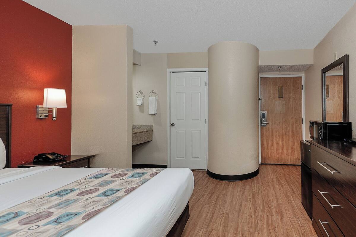 A hotel room with a bed, a desk with a lamp, a telephone, a microwave, a wooden dresser, and a bathroom with a sink and towel rack.