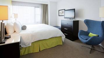 A hotel room with a bed, a TV, a desk with a lamp, a chair, and a window with curtains.