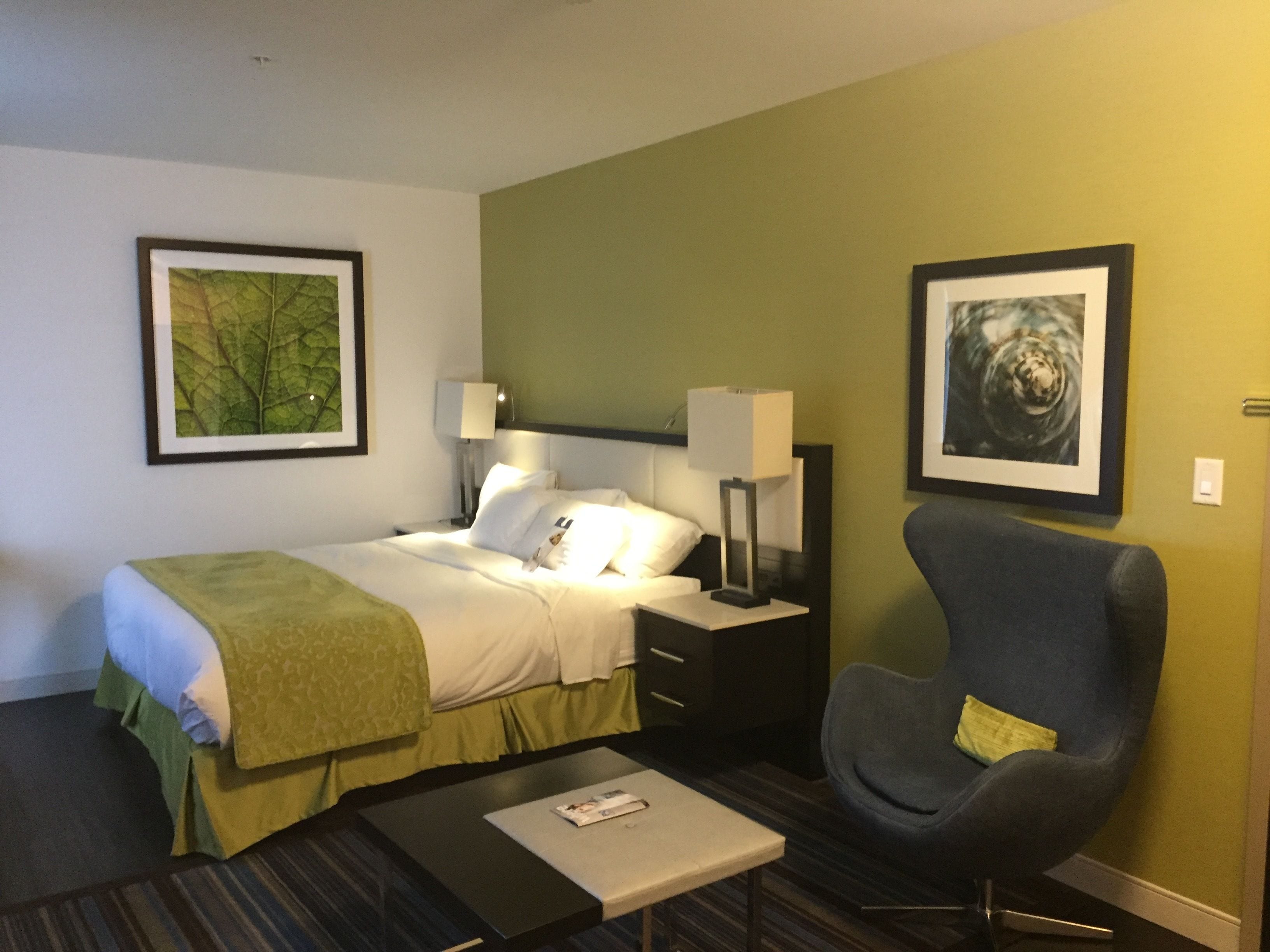 Habitación de hotel con cama, una silla, una mesita de noche, lámpara y dos cuadros en la pared.