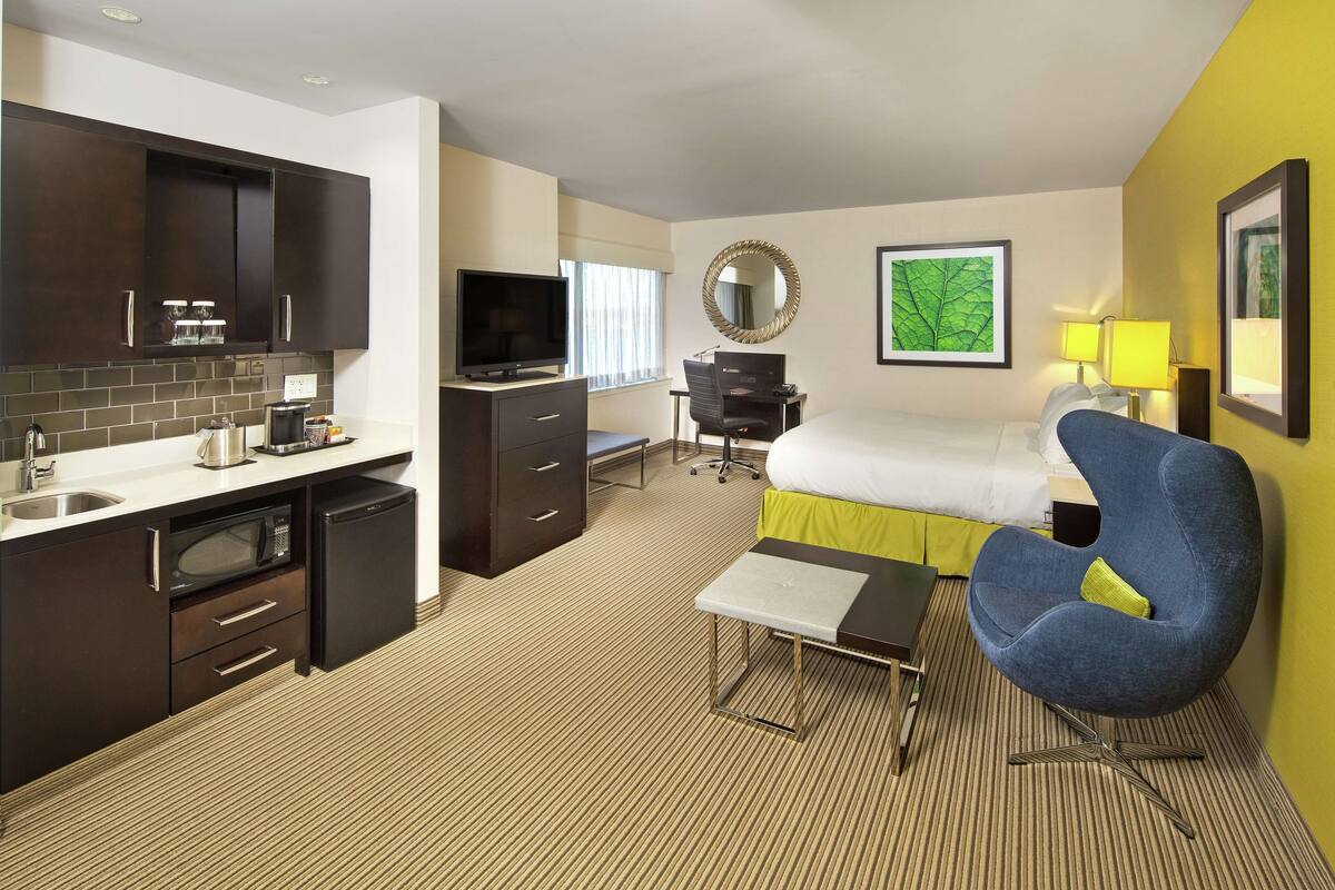 A hotel room with a kitchenette, a bed, a desk, a chair, and a TV.