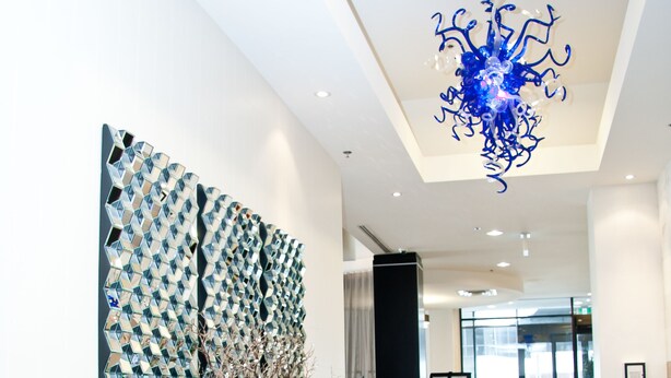 A modern lobby with a wooden reception desk, a blue chandelier, and a geometric wall art piece.
