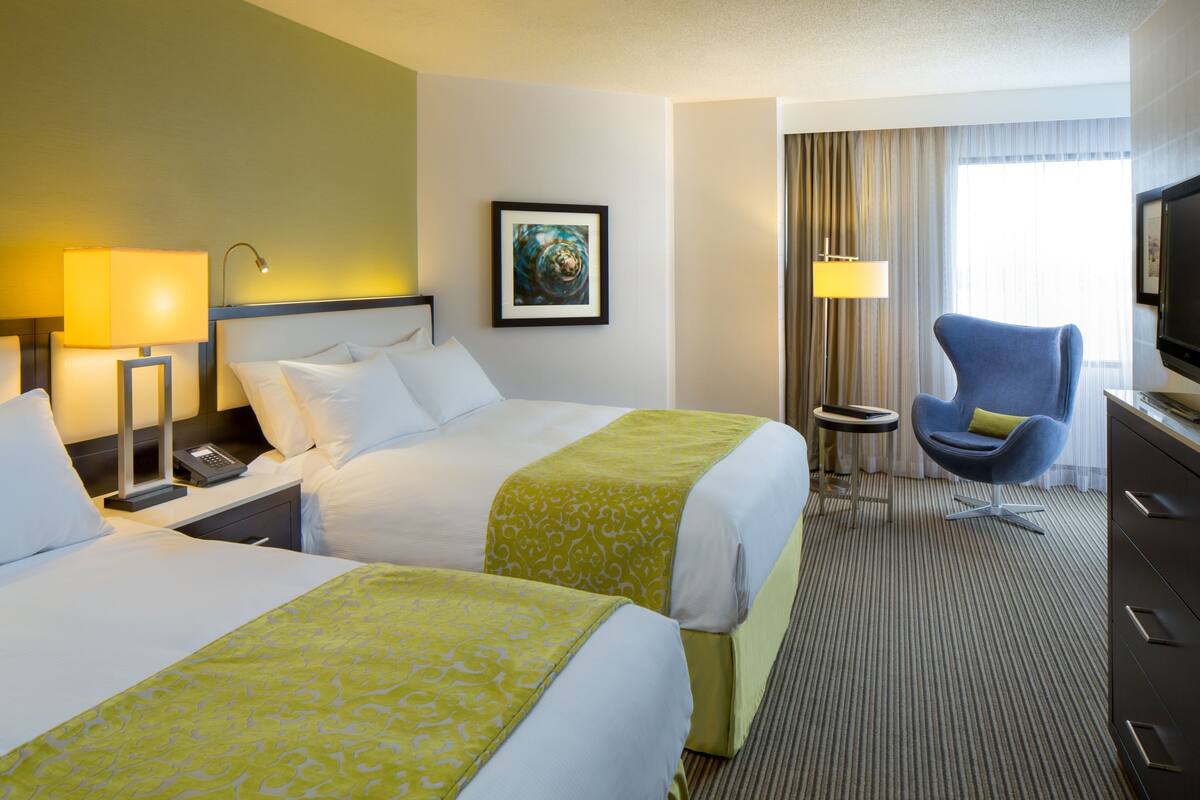 A hotel room with two beds, a desk with a lamp, a chair, a television, and a painting on the wall.