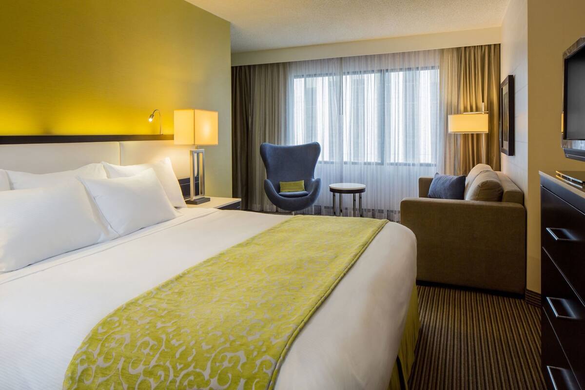 A hotel room with a large bed, a desk, a chair, a sofa, a TV, and a window with curtains.