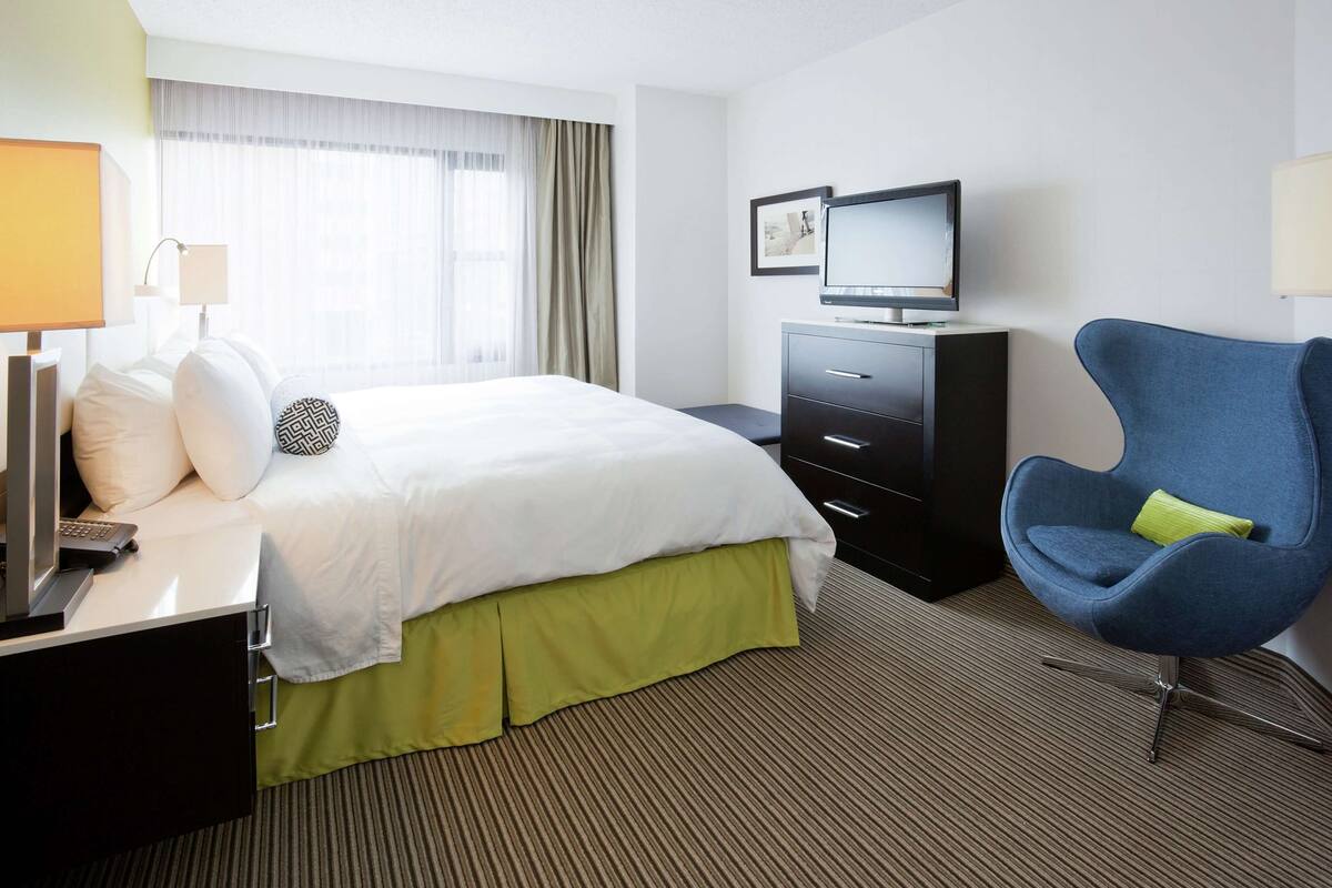 A hotel room with a bed, a TV, a desk with a lamp, a chair, and a window with curtains.