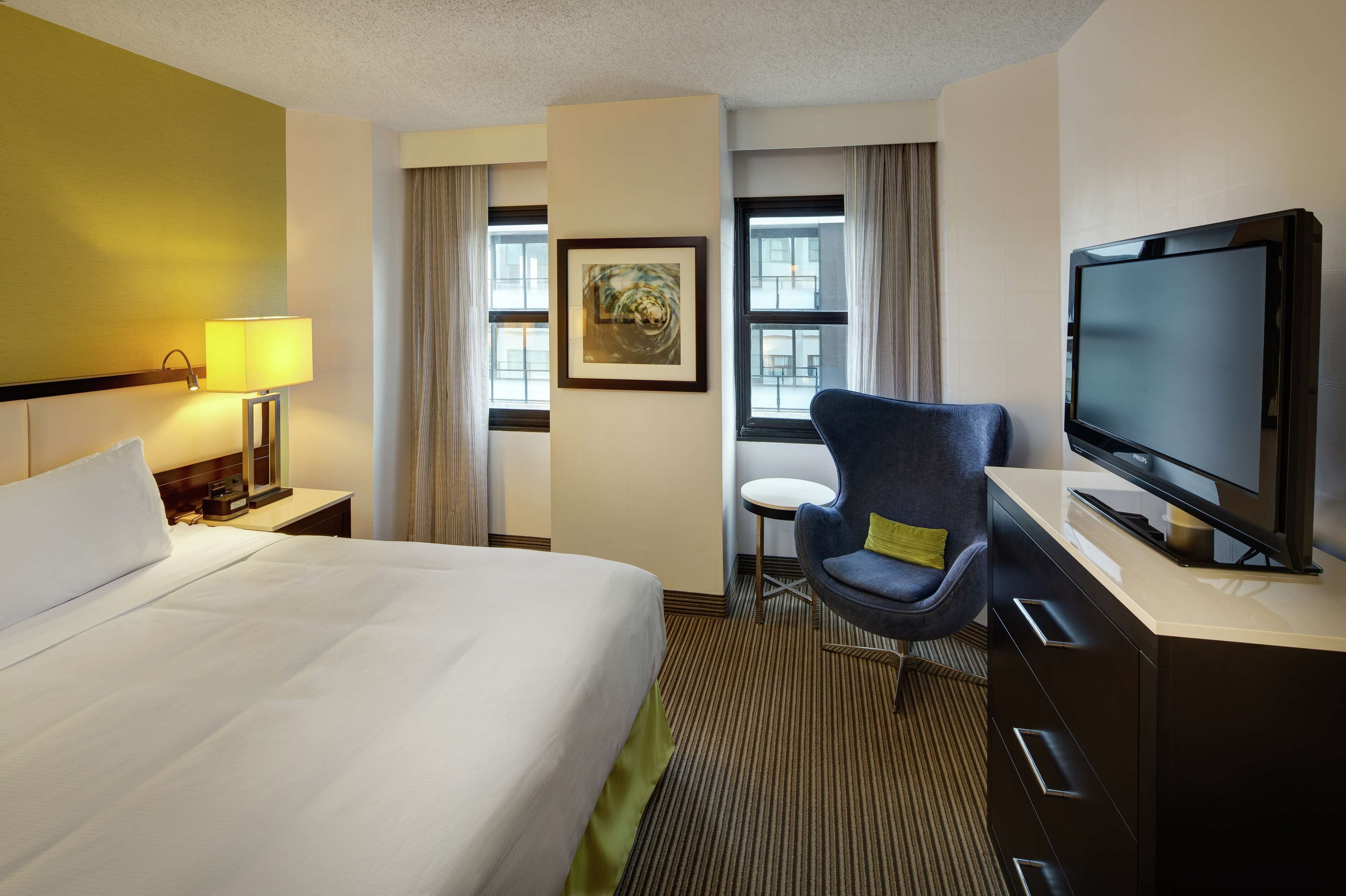 A hotel room with a bed, a desk with a television, a chair, a lamp, and a window with curtains.