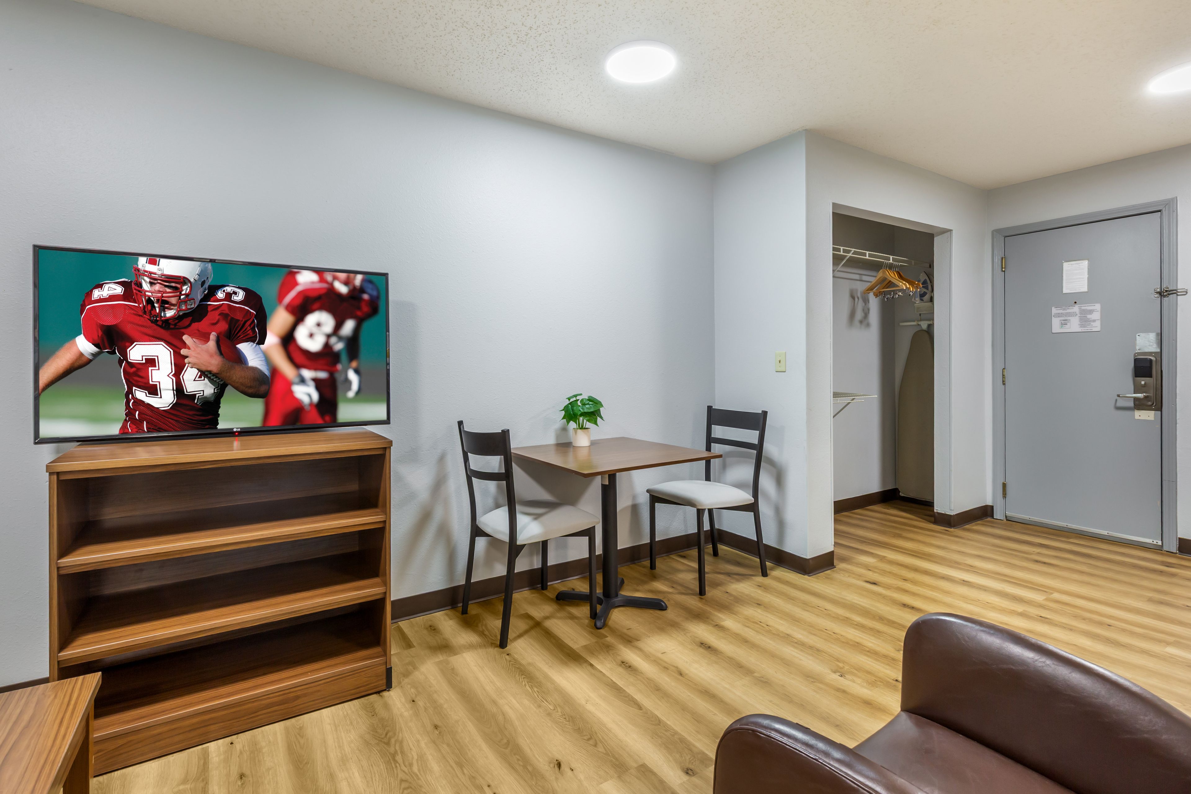 A room with a TV displaying sports players, a wooden shelf, a table with chairs, and a coat rack.