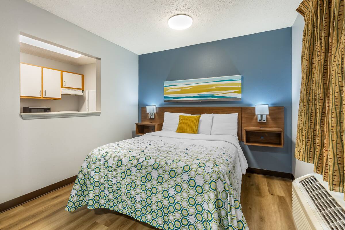A hotel room with a bed, a nightstand with a lamp, a wall-mounted artwork, and a window with curtains.