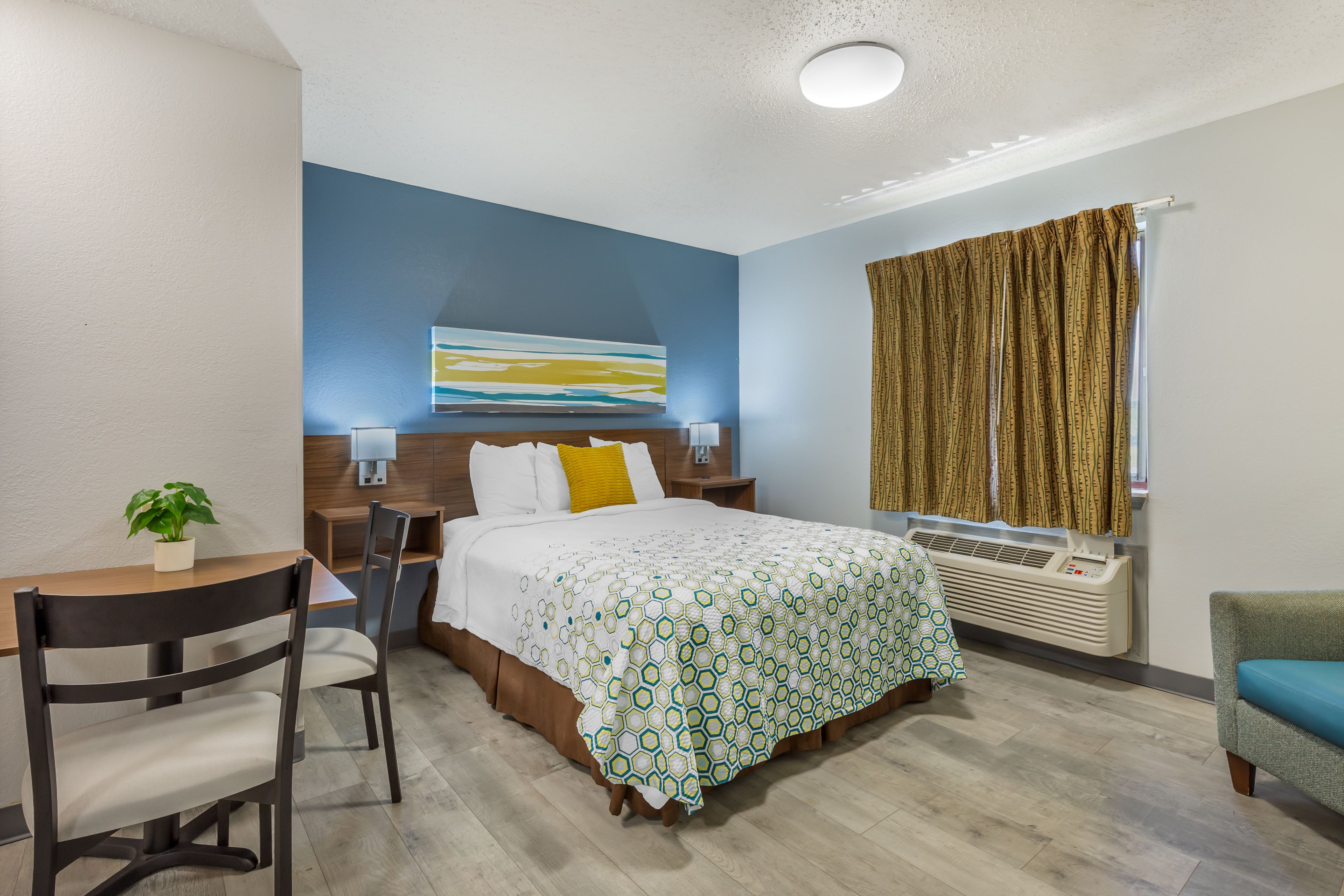 A hotel room with a bed, a desk with a chair, a potted plant, a window with curtains, and a wall-mounted air conditioner.