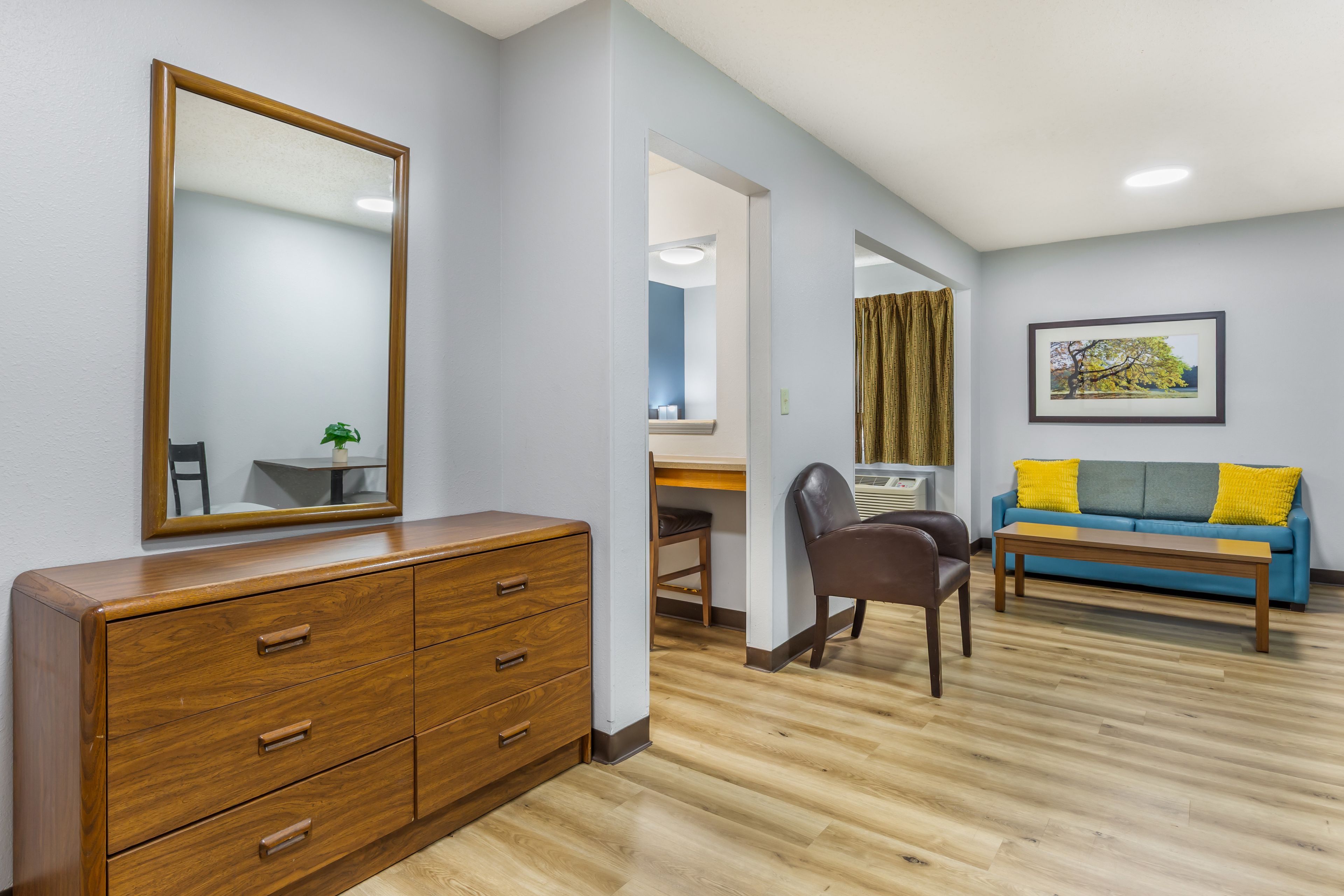 A room with a wooden dresser, a desk, a chair, a blue sofa with yellow cushions, and a framed picture on the wall.