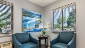 A room with two blue armchairs, a small table with a lamp, a painting, and a window with blinds.