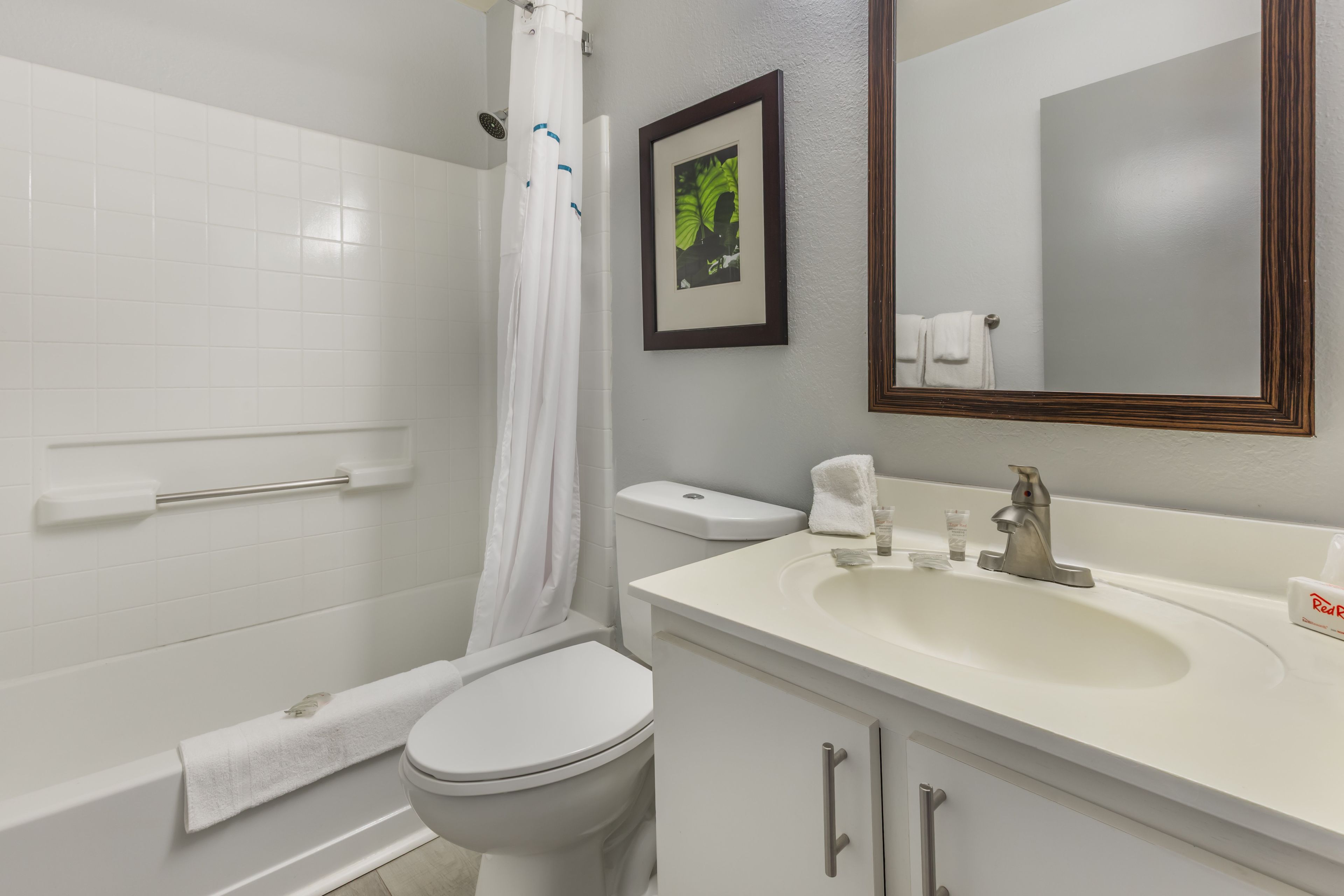 Ein Badezimmer mit Toilette, einer Badewanne mit weißem Duschvorhang, einem Waschbecken mit Spiegel und einem gerahmten Bild an der Wand.