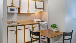 A compact kitchen with white cabinets, a microwave, a stove, a sink, and a dining table with two chairs. - HomeTowne Studios & Suites by Red Roof Greensboro (Greensboro)