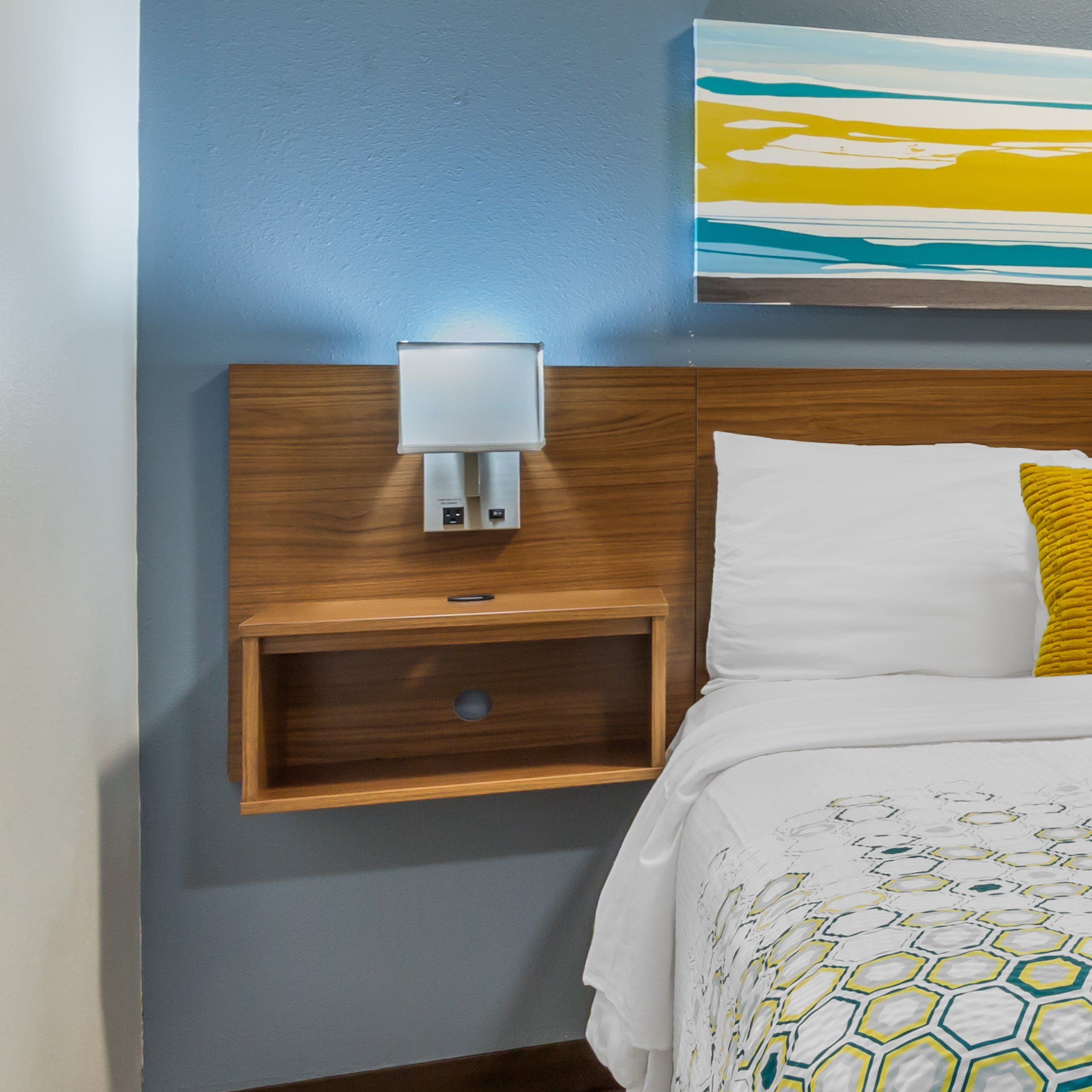 A hotel room with a wooden headboard, a bedside table with a lamp, a white bed with a yellow pillow, and a painting above the bed.