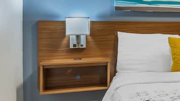 A hotel room with a wooden headboard, a bedside table with a lamp, a white bed with a yellow pillow, and a painting above the bed.