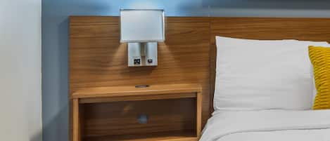 Habitación de hotel con cabecera de madera, una mesita de noche con lámpara, cama blanca con almohada amarilla y un cuadro sobre la cama.