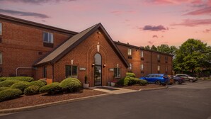 A brick building with a lobby entrance, a parking lot with several cars, and well-maintained landscaping. - HomeTowne Studios & Suites by Red Roof Greensboro (Greensboro)