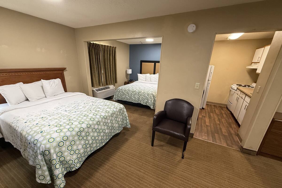 A hotel room with two beds, a chair, a kitchenette, and a bathroom visible through an open door.