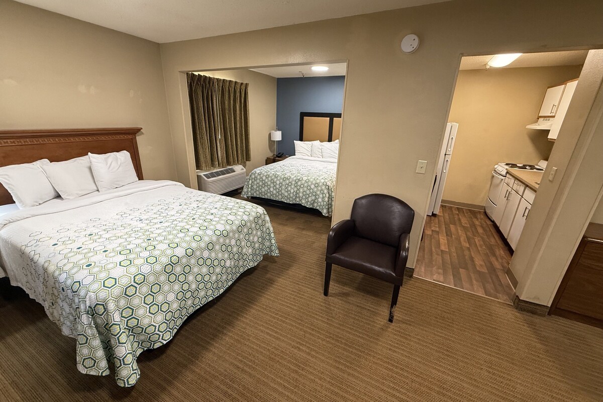 Une chambre d’hôtel avec deux lits, une chaise, un coin cuisine et une salle de bain visible à travers une porte ouverte.