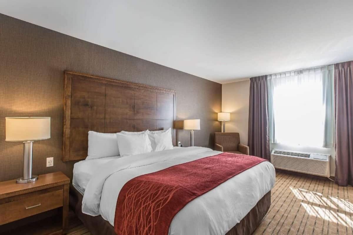 A hotel room with a large bed, two bedside lamps, a chair, a window with curtains, and an air conditioning unit.
