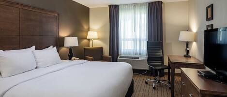 Habitación de hotel con una cama grande, un escritorio con televisor, una silla, una lámpara y una ventana con cortinas.